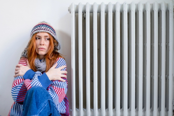 woman feeling cold wrapped in winter clothes beside home heating radiator due to oil fuel running low